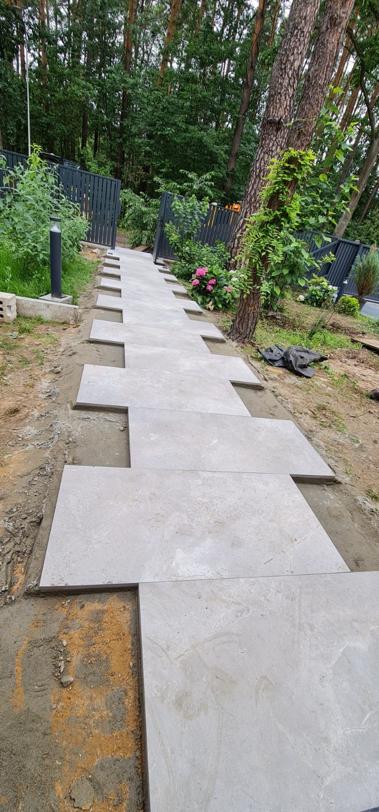 Kładzenie jasnych, prostokątnych płyt chodnikowych na ścieżce w ogrodzie, widoczne przerwy między płytami wypełnione piaskiem, w tle drzewa i ogrodzenie.