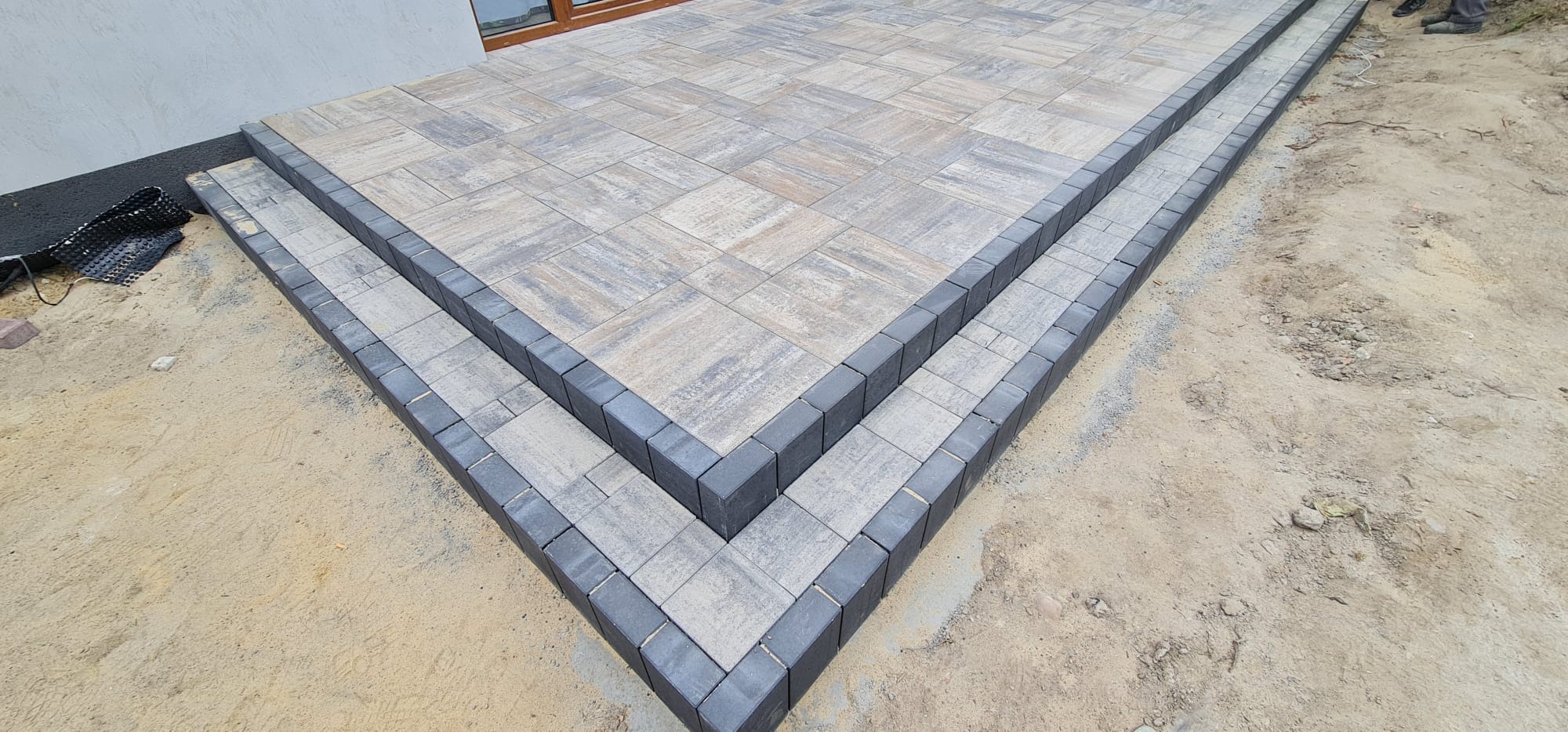 Schody z kostki brukowej w odcieniach szarości, z ciemniejszym obramowaniem, prowadzące do jasnej nawierzchni z kostki. Widoczny fragment elewacji budynku w tle i piasek wokół.