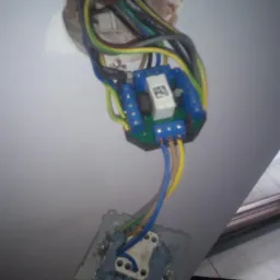 Otwarta puszka elektryczna w ścianie z podłączonym modułem sterującym i widocznym gniazdkiem, przewody w kolorach żółtym, niebieskim, zielonym i brązowym.