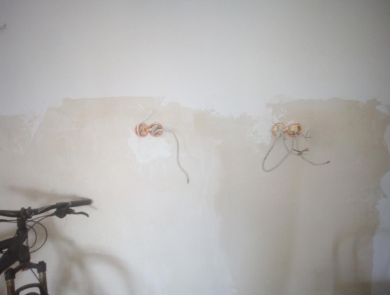Dwa gniazda elektryczne w ścianie po tynkowaniu, wystające przewody, fragment roweru w lewym dolnym rogu.