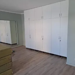 Białe szafy w zabudowie, z metalowymi uchwytami, na tle szarej ściany i drewnopodobnej podłogi, z odbiciem w lustrze i kartonami na pierwszym planie.