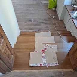 Wykończone schody z płytek imitujących drewno, zabezpieczone kartonem na czas remontu, widoczne akcesoria i narzędzia budowlane w tle.