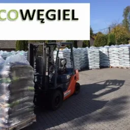 Pomarańczowy wózek widłowy Toyota podnosi paletę z workami owiniętymi folią stretch, w tle stosy palet z workami i budynki.