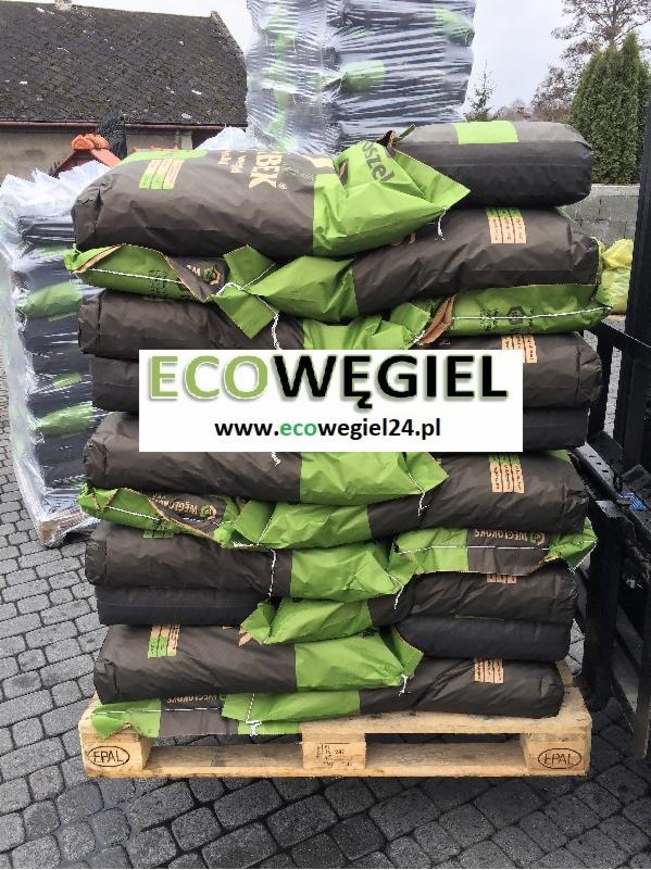 Paleta z workami opału w kolorze grafitowo-zielonym, ustawiona na brukowanym podłożu, z widocznym budynkiem w tle.