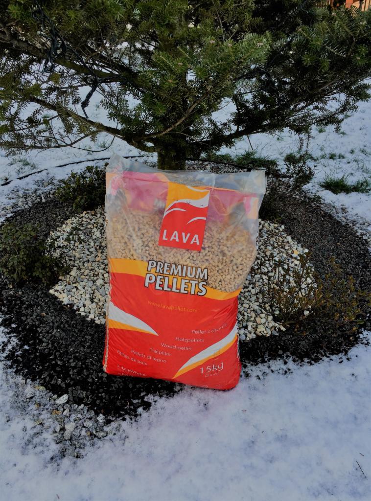 Worek z pelletem drzewnym marki Lava, ustawiony na tle kamiennego klombu pod drzewem iglastym, z widocznym śniegiem w tle.