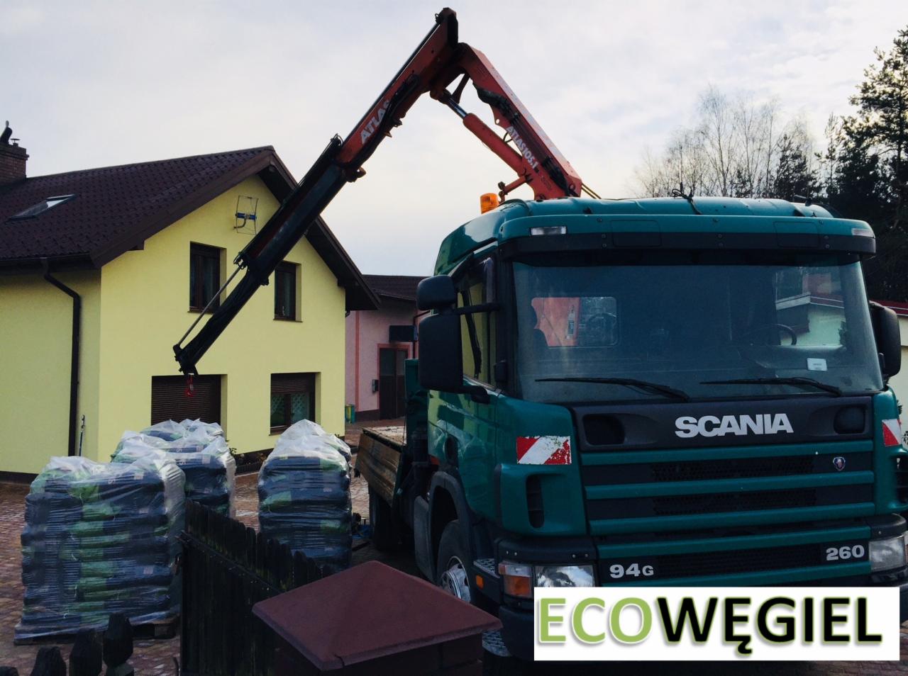 Zielona ciężarówka Scania z dźwigiem Atlas dostarcza zapakowany ekogroszek przed żółty dom.