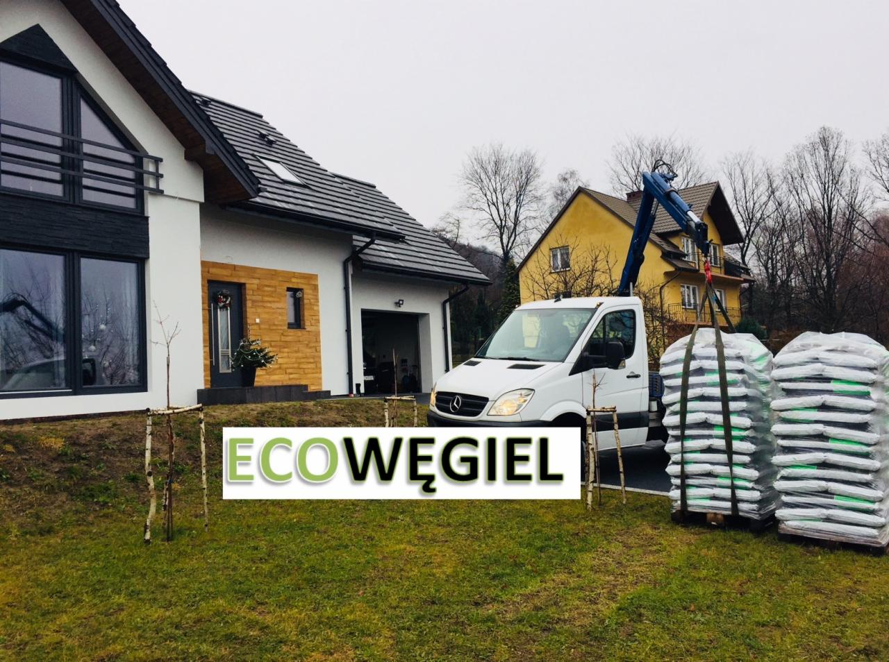 Dostawa workowanego opału, prawdopodobnie ekogroszku, dźwigiem samochodowym na posesję z nowoczesnym domem jednorodzinnym.