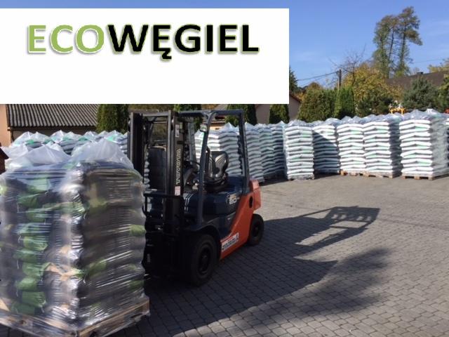 Pomarańczowy wózek widłowy Toyota podnosi paletę z workami owiniętymi folią stretch, w tle stosy palet z workami i budynki.