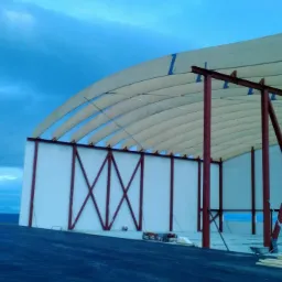 Konstrukcja hali z białymi ścianami i łukowym dachem z belek, widoczne czerwone stalowe elementy konstrukcyjne, w tle krajobraz z morzem i zachmurzonym niebem.