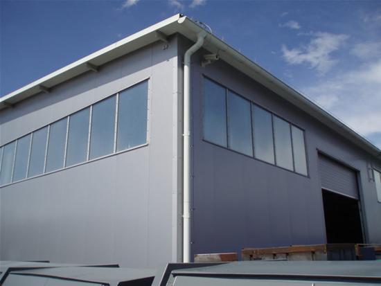 Szary budynek przemysłowy z panelami, widoczne okna w rzędzie, brama garażowa częściowo otwarta oraz rynna na krawędzi dachu.