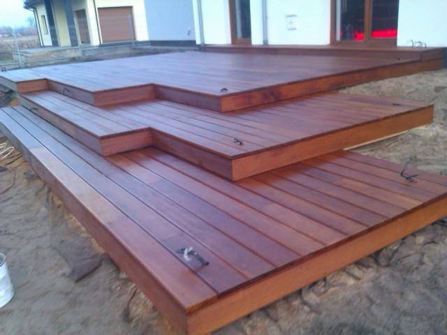 Nowoczesny drewniany taras z wyraźnymi schodkowymi poziomami, świeżo po impregnacji, z widocznym wiaderkiem farby i elementami mocującymi w tle domu jednorodzinnego.