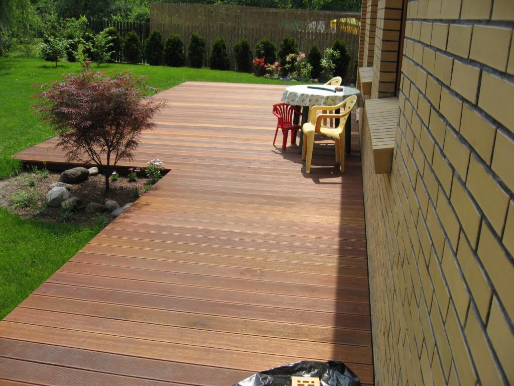 Drewniany taras przydomowy z desek kompozytowych, z małym klonem palmowym, stołem i krzesłami na tle zadbanego ogrodu i ściany z żółtej cegły.