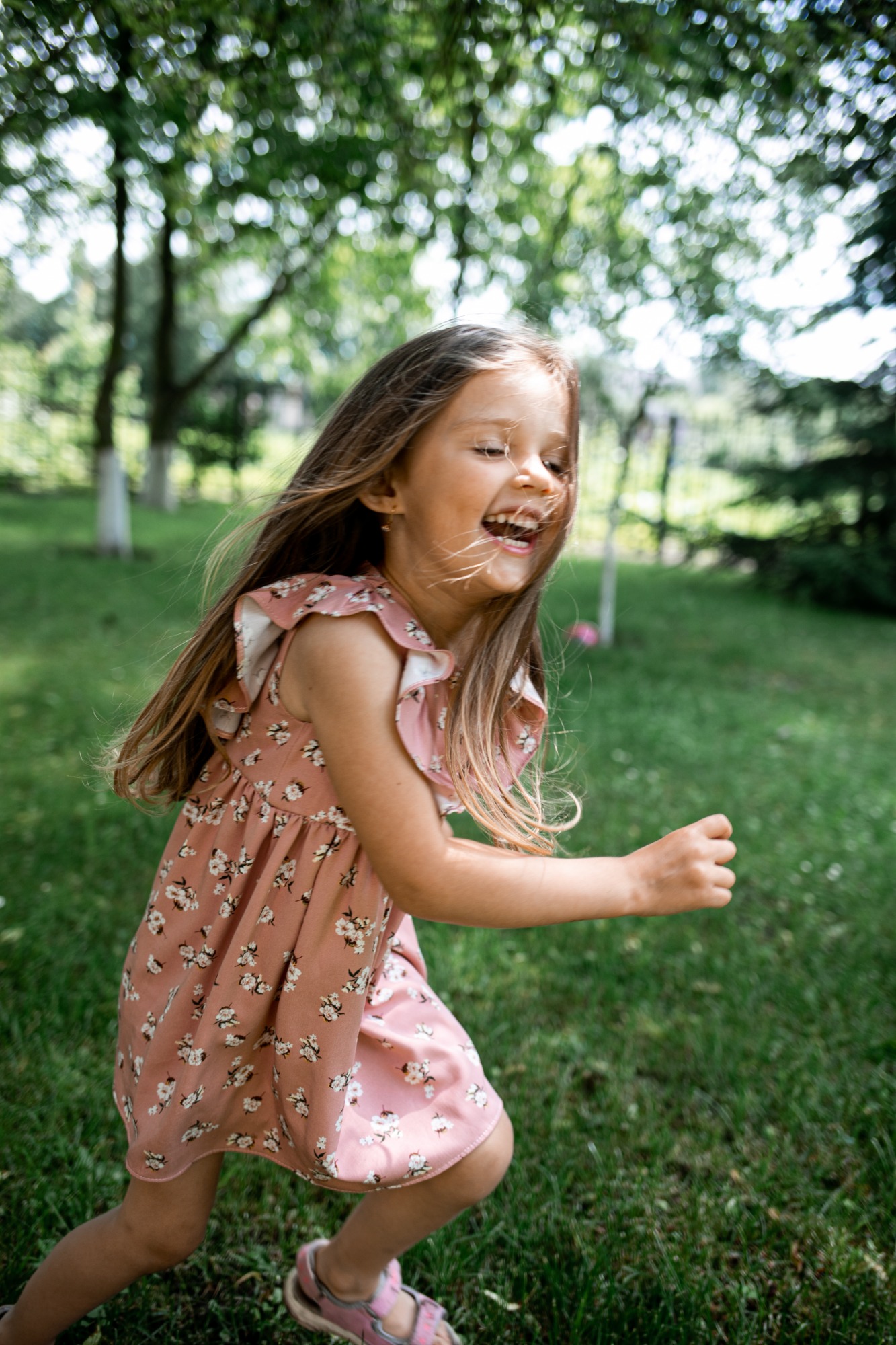 Uśmiechnięta dziewczynka w kwiecistej, różowej sukience z falbankami biegnie po trawie w słoneczny dzień.
