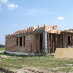 Wznoszenie muru z czerwonej cegły na fundamencie, widoczne drewniane szalunki i podpory, zbrojenia stalowe leżące na trawie, w tle zieleń drzew.