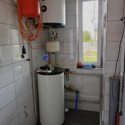Usługi Instalacyjne Zbigniew Kryszkiewicz - Bojler i instalacja CO w łazience z oknem, Łowicz. Widoczne rury, zawory i zbiornik wyrównawczy. Białe płytki na ścianach, szara podłoga.