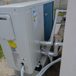 Usługi Instalacyjne Zbigniew Kryszkiewicz - Biała pompa ciepła zamontowana na zewnątrz budynku, z widocznymi rurami w srebrnej izolacji i przewodami. W tle betonowy płot i budynek.