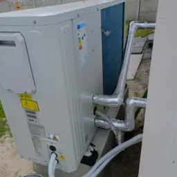 Biała pompa ciepła zamontowana na zewnątrz budynku, z widocznymi rurami w srebrnej izolacji i przewodami. W tle betonowy płot i budynek.