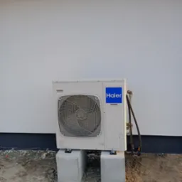 Biała jednostka zewnętrzna klimatyzacji Haier, zamontowana na betonowych podstawach przy ścianie budynku. Widoczne rury i przewody. Ujęcie z poziomu gruntu.