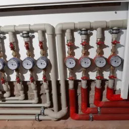 Rozdzielacz instalacji wodnej z czterema wodomierzami po lewej i czterema po prawej stronie, rury z czerwonym ociepleniem po prawej, całość umieszczona w białej szafce.