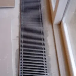 Grzejnik kanałowy zamontowany w podłodze przy oknie, widoczne niedoskonałości wykończenia ściany i podłogi.