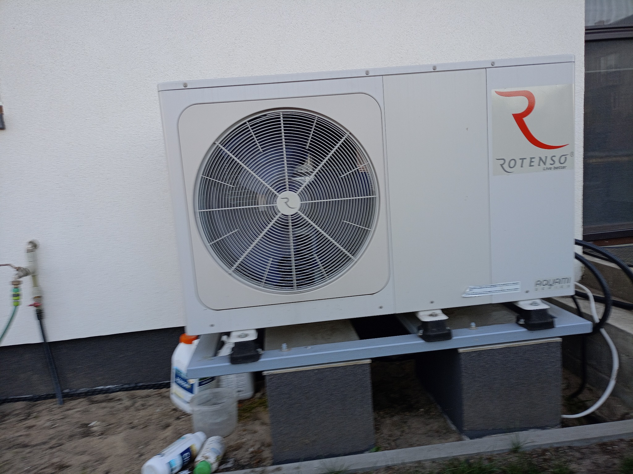 Biała pompa ciepła Rotenso Aquami Series, zamontowana na szarej konstrukcji wsporczej przy ścianie budynku. Widoczne elementy instalacji i akcesoria montażowe.