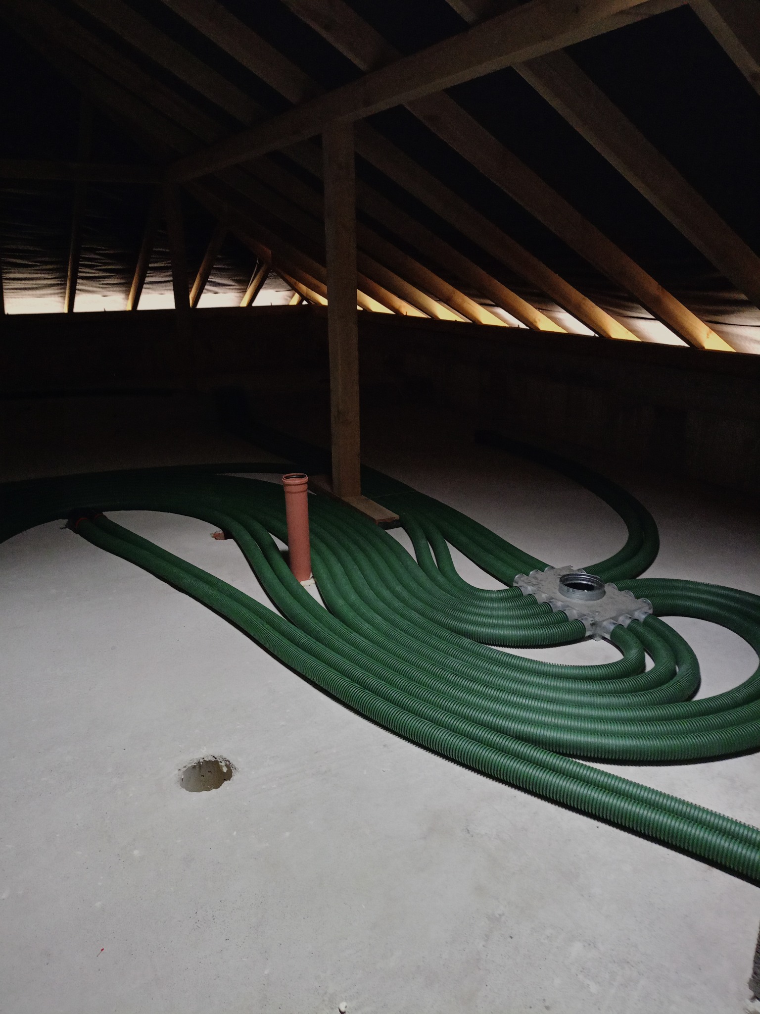 Zielone rury wentylacyjne ułożone na betonowej podłodze poddasza, widoczne drewniane elementy konstrukcji dachu i pionowa rura wentylacyjna. Instalacja w trakcie montażu.