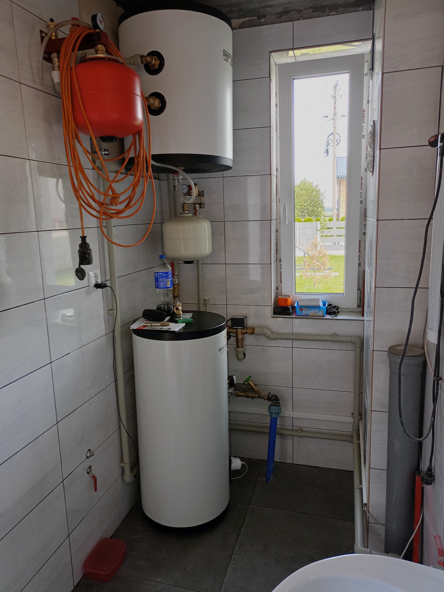 Bojler i instalacja CO w łazience z oknem, Łowicz. Widoczne rury, zawory i zbiornik wyrównawczy. Białe płytki na ścianach, szara podłoga.