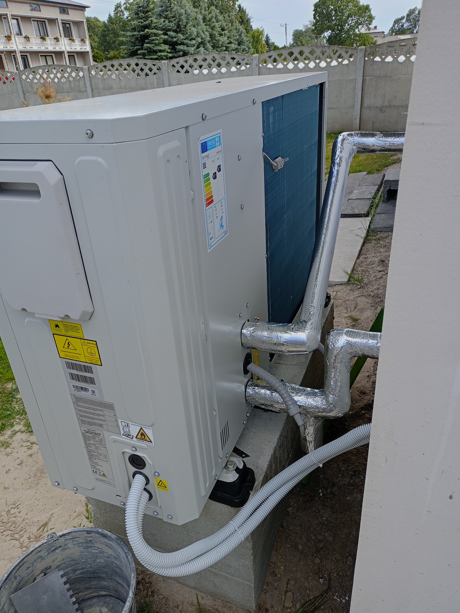 Biała pompa ciepła zamontowana na zewnątrz budynku, z widocznymi rurami w srebrnej izolacji i przewodami. W tle betonowy płot i budynek.