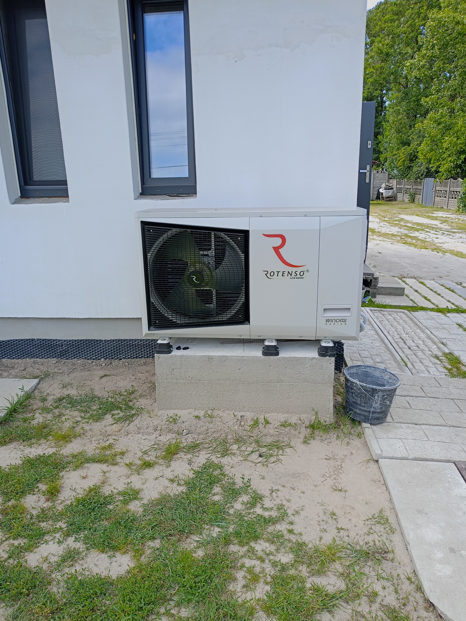 Biała pompa ciepła Rotenso Windmi Series zamontowana na betonowym fundamencie przy ścianie budynku z dwoma oknami. Obok stoi wiadro.