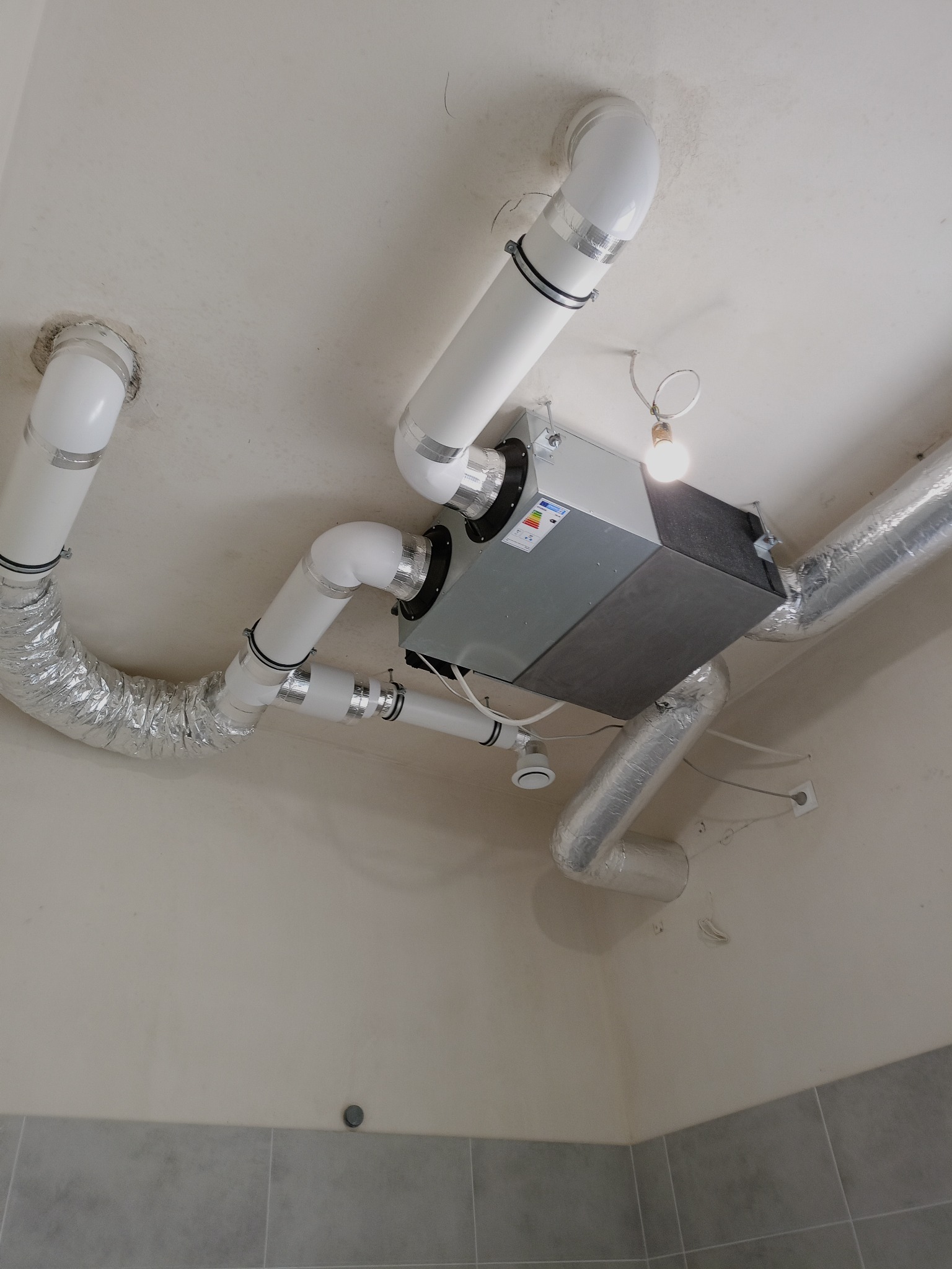 Szara centrala wentylacyjna z białymi i aluminiowymi rurami przymocowana do sufitu. Widoczne elementy instalacji i oświetlenie. Ujęcie z dołu.