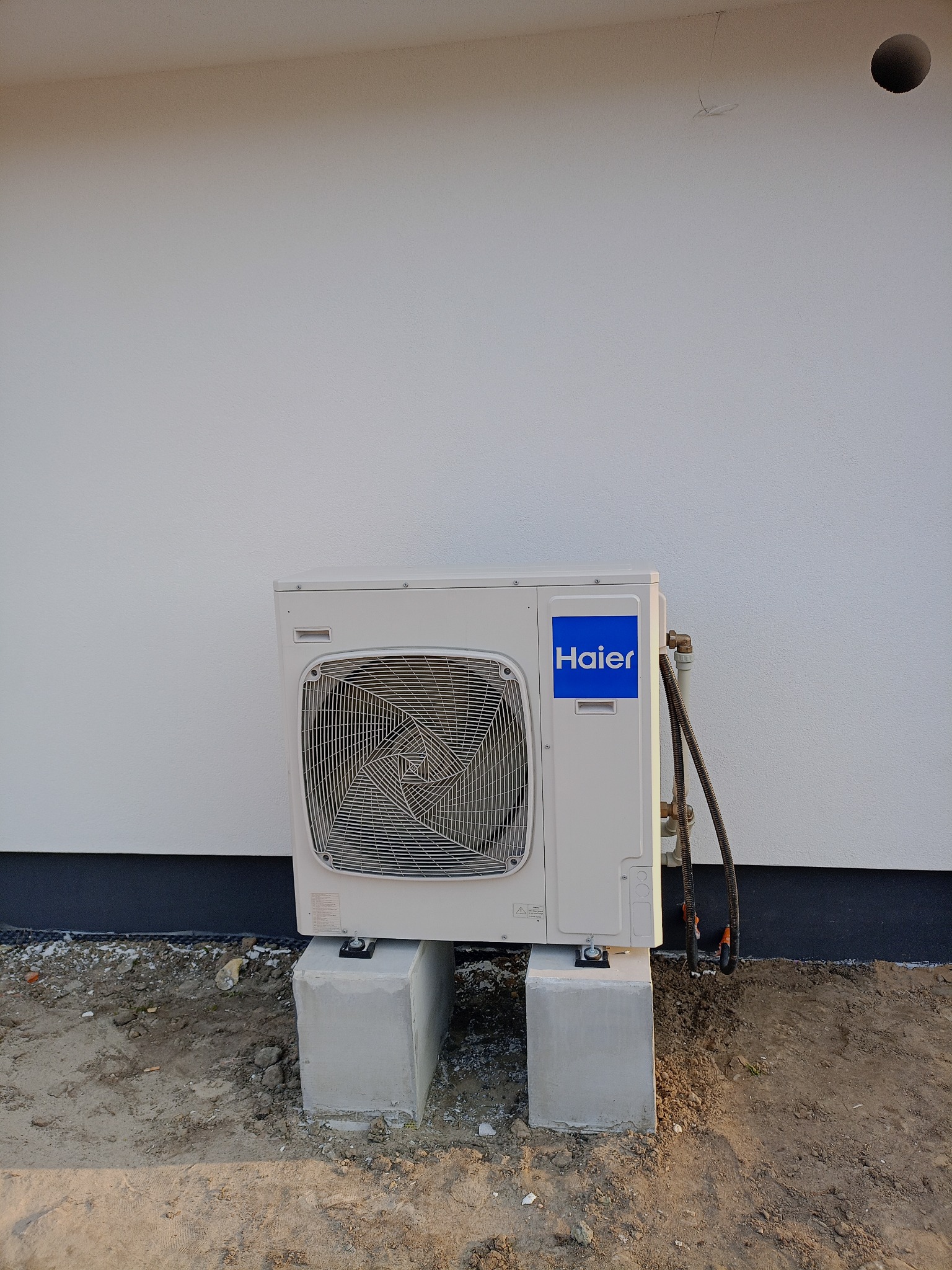 Biała jednostka zewnętrzna klimatyzacji Haier, zamontowana na betonowych podstawach przy ścianie budynku. Widoczne rury i przewody. Ujęcie z poziomu gruntu.