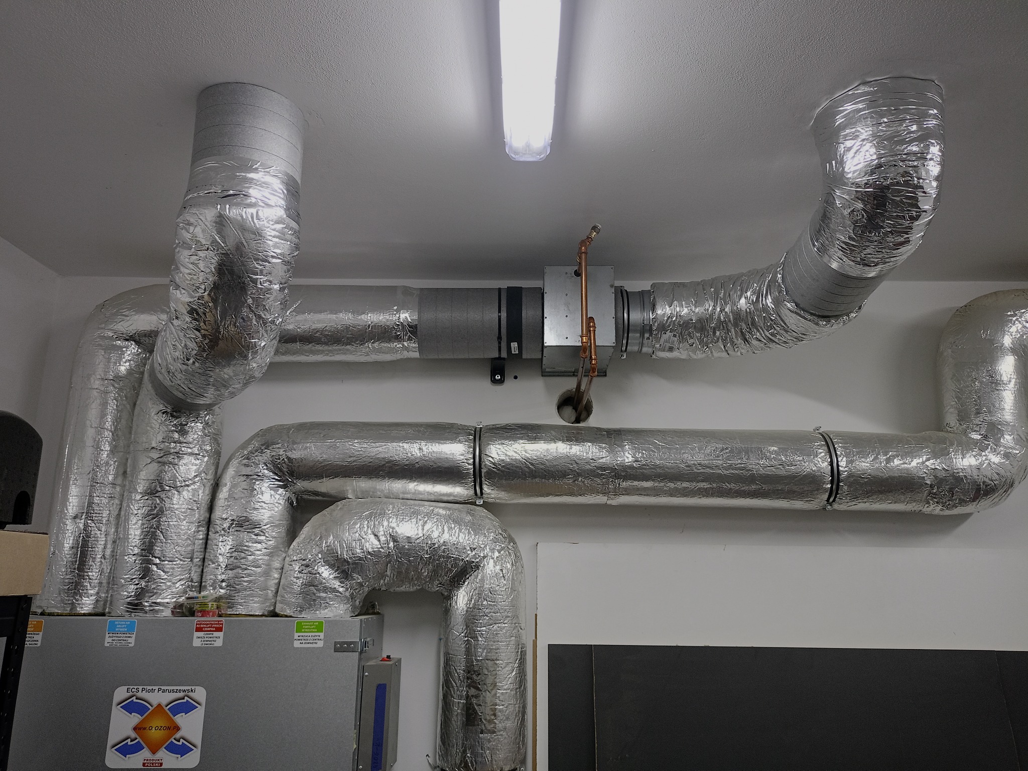 Kompleksowa instalacja wentylacyjna z rurami w srebrnej izolacji, widoczne elementy centrali wentylacyjnej i oświetlenie sufitowe.