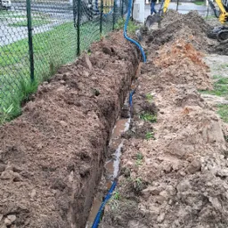 Głęboki wykop w ziemi z niebieską rurą karbowaną na dnie, żółta koparka w tle, widoczne budynki mieszkalne i ogrodzenie z siatki.