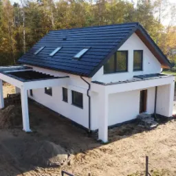 Nowoczesny dom jednorodzinny z ciemną dachówką i białymi ścianami, widok z drona na plac budowy z widocznymi stertami piasku i sąsiednim domem w stanie surowym.