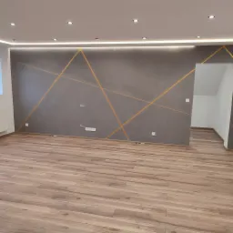 Szara ściana z geometrycznym wzorem w odcieniach złota, oświetlenie sufitowe LED, panele podłogowe imitujące drewno, widoczne gniazdka elektryczne i włączniki, minimalistyczne wykończenie wnętrza.