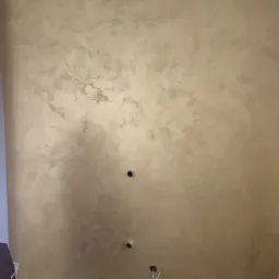 Ściana w odcieniach złota z dekoracyjną strukturą, widoczne otwory na gniazdka elektryczne i fragment podłogi w trakcie prac wykończeniowych.