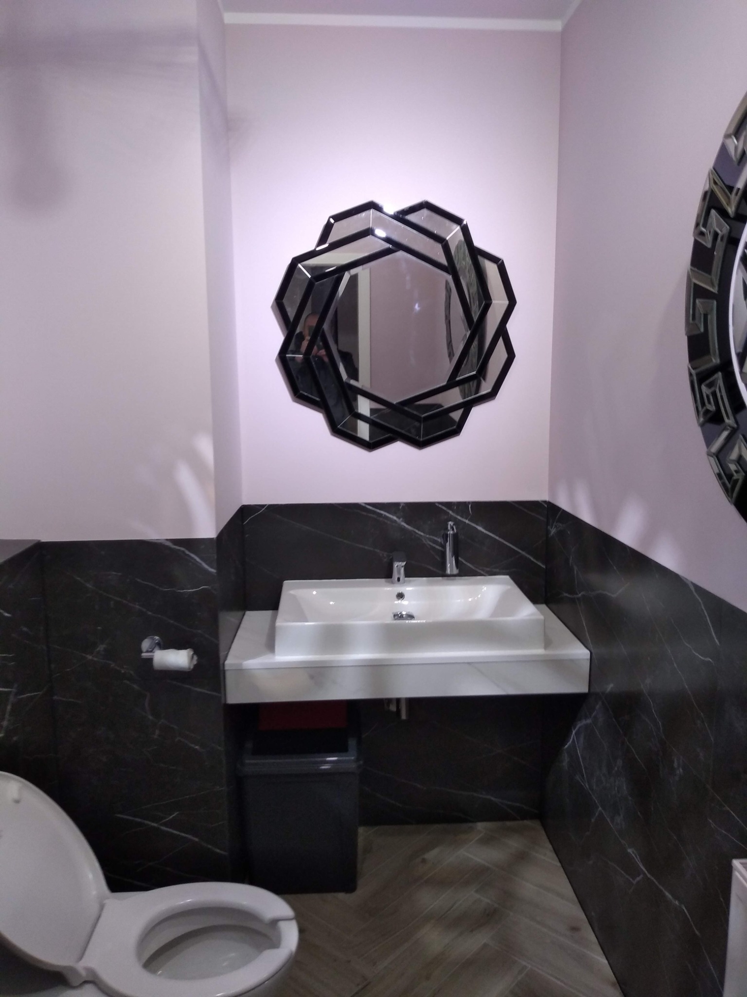 Nowoczesna łazienka z umywalką nablatową, geometrycznym lustrem i czarnymi marmurowymi płytkami do połowy ściany.