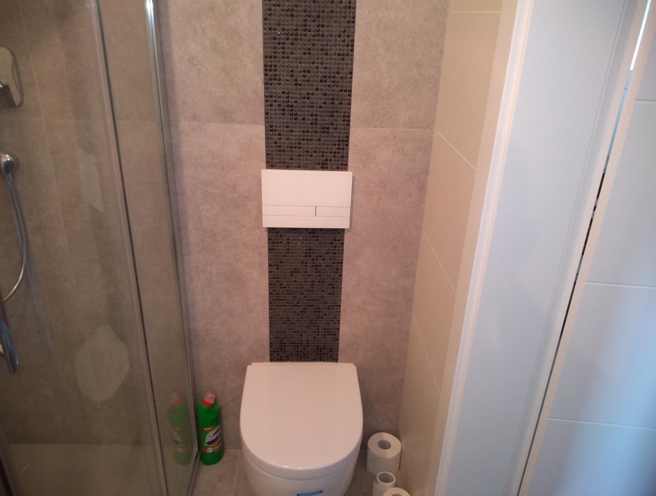 Toaleta w łazience z szarymi płytkami i mozaiką, widoczna kabina prysznicowa z butelką środka czyszczącego i rolki papieru toaletowego.