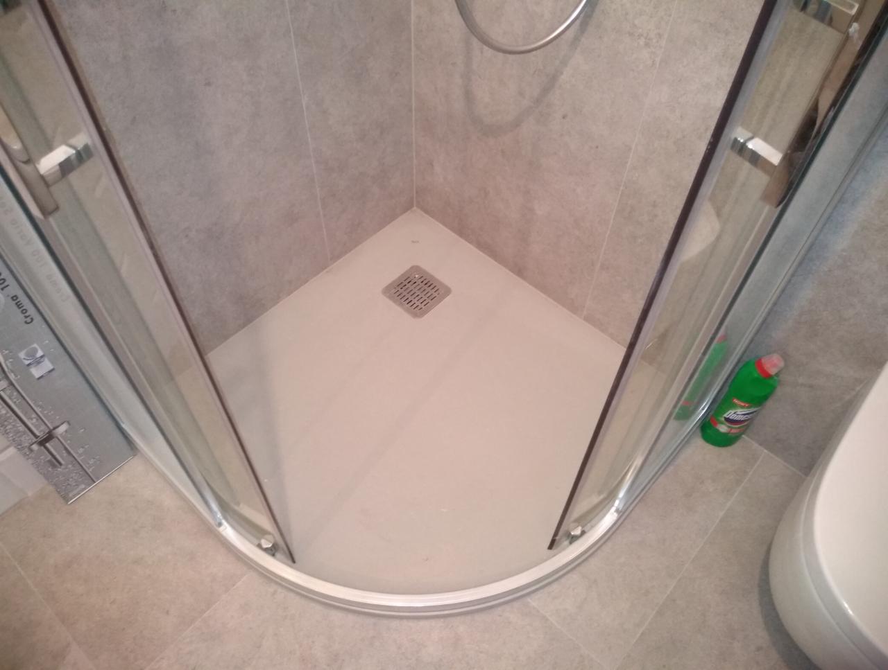 Wykończony narożny brodzik z odpływem, kabina prysznicowa z chromowanymi profilami, szare płytki na ścianach i podłodze, butelka płynu do czyszczenia w rogu.