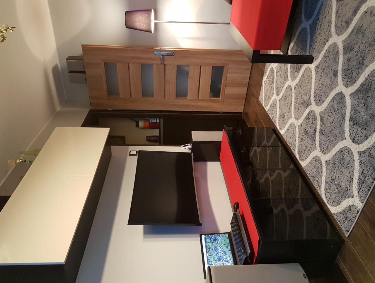 Nowoczesny salon z białymi szafkami wiszącymi, czarną szafką RTV z czerwonym akcentem, drewnianymi drzwiami z przeszkleniami oraz szarym dywanem we wzory.