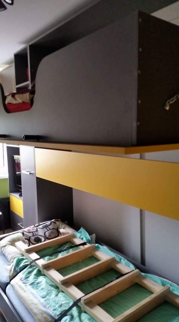 Wysokie łóżko z szarą szafą na górze i żółtym biurkiem pod spodem, drabinka na łóżku, widok z dołu.