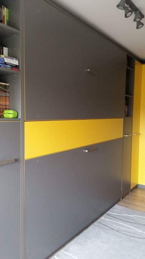Szara zabudowa ścienna z żółtym pasem, wbudowana szafa z półkami, minimalistyczne uchwyty, oświetlenie sufitowe.