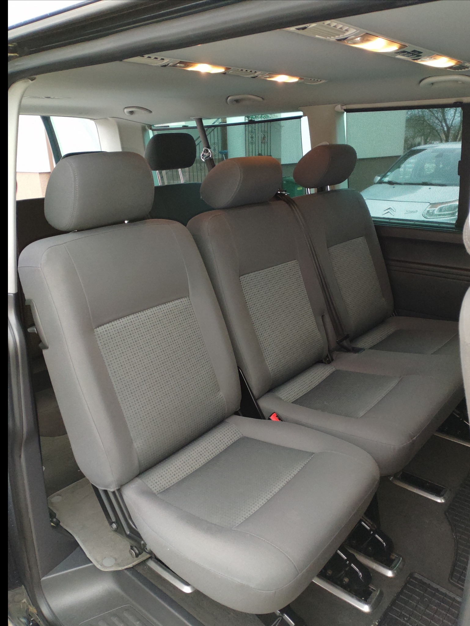 Wnętrze busa z trzema rzędami szarych siedzeń z zagłówkami, widoczne oświetlenie sufitowe i fragment samochodu za oknem.