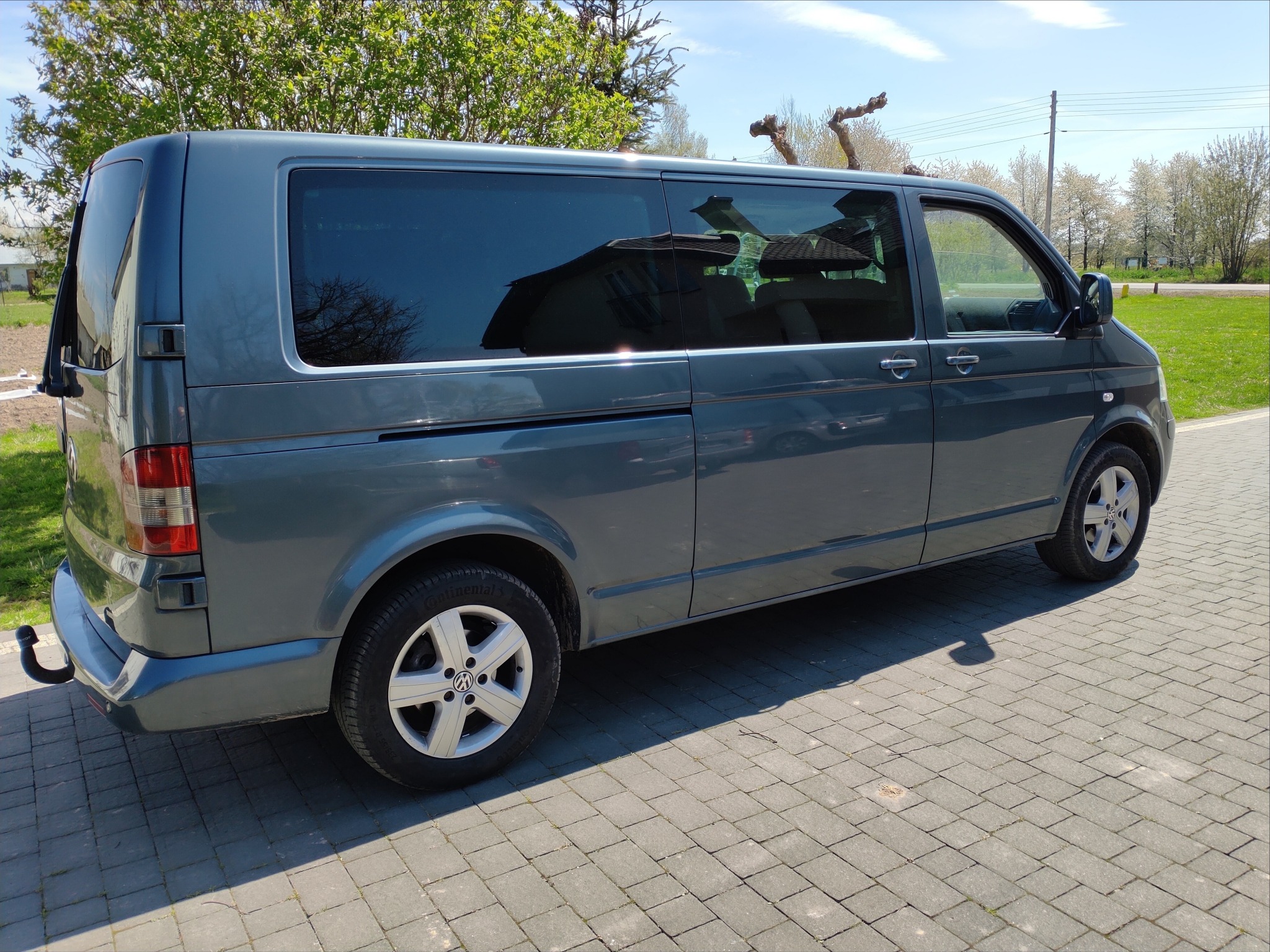 Szary Volkswagen Transporter T5 z przyciemnianymi szybami, zaparkowany na kostce brukowej w słoneczny dzień, z widocznym hakiem holowniczym.