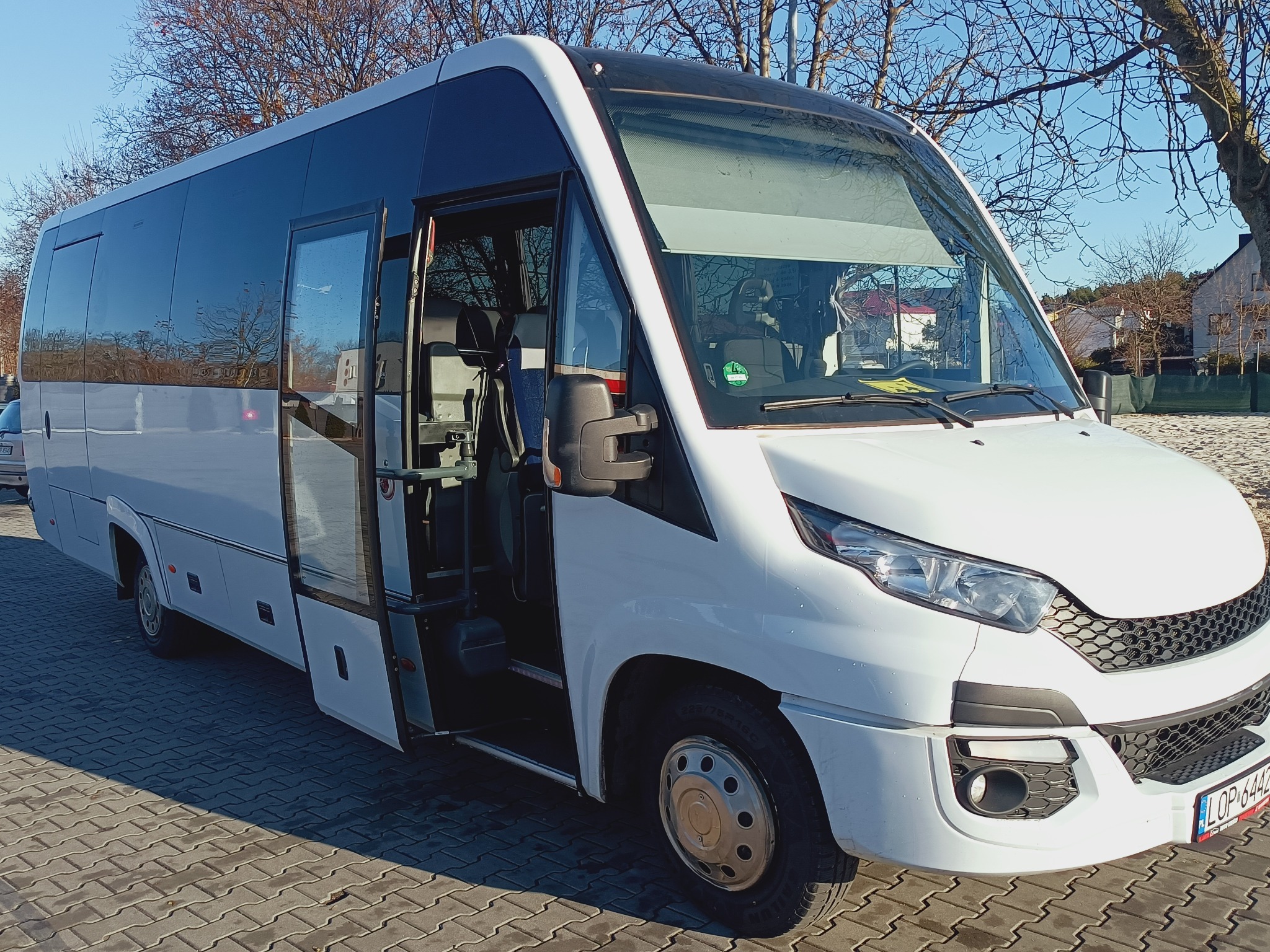 Biały minibus Iveco z otwartymi drzwiami wejściowymi, zaparkowany na brukowanej powierzchni, widoczne drzewa bez liści w tle.