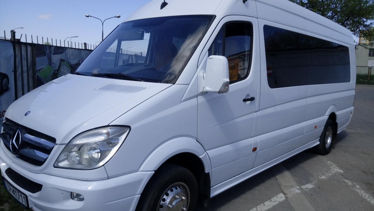 Biały bus Mercedes-Benz Sprinter z przyciemnianymi szybami, zaparkowany na betonowym parkingu, widok z przodu i boku.