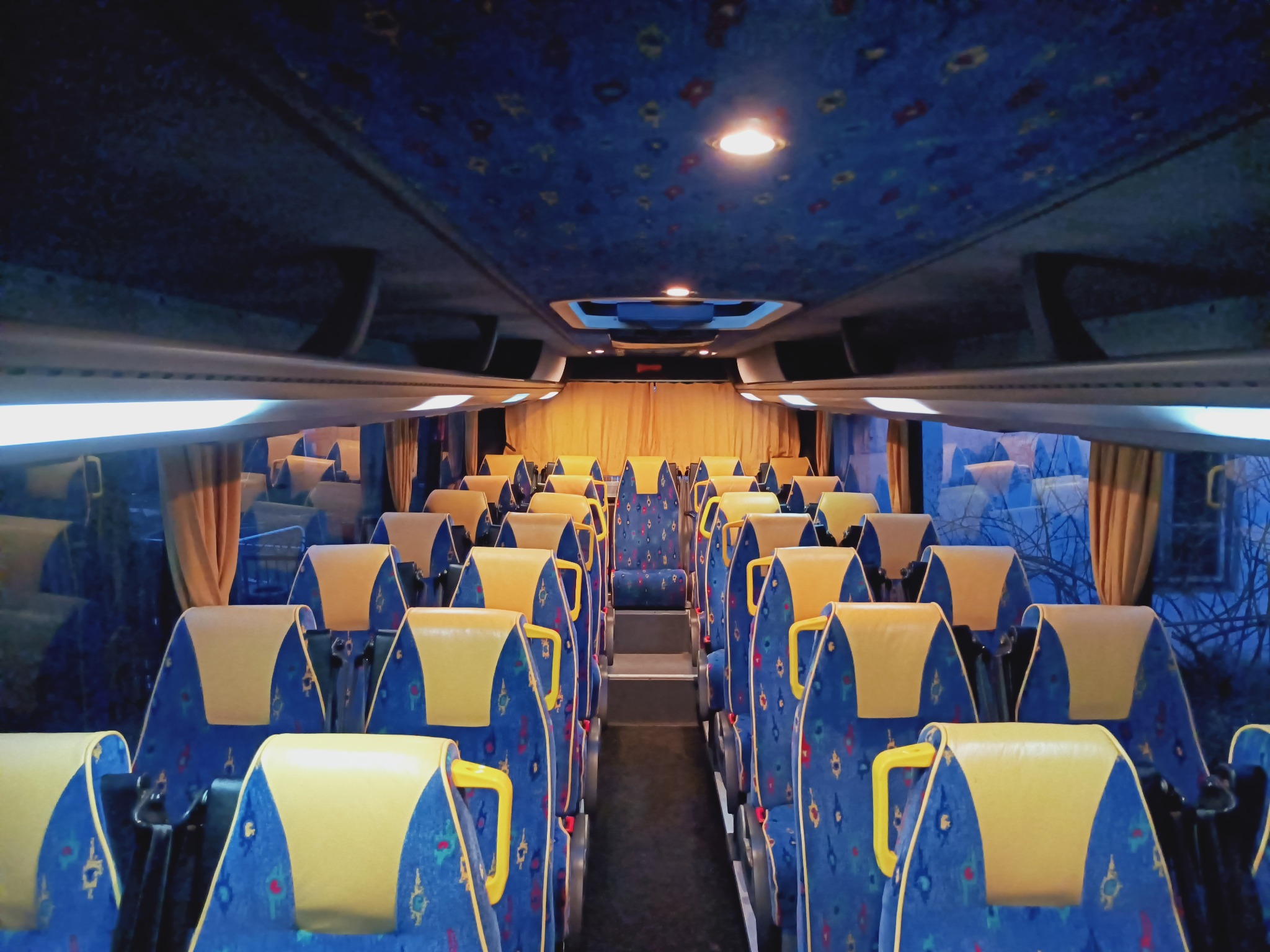 Wnętrze autobusu turystycznego z rzędami niebieskich siedzeń z żółtymi zagłówkami, widok wzdłuż korytarza, zasłonięte okna w tle.