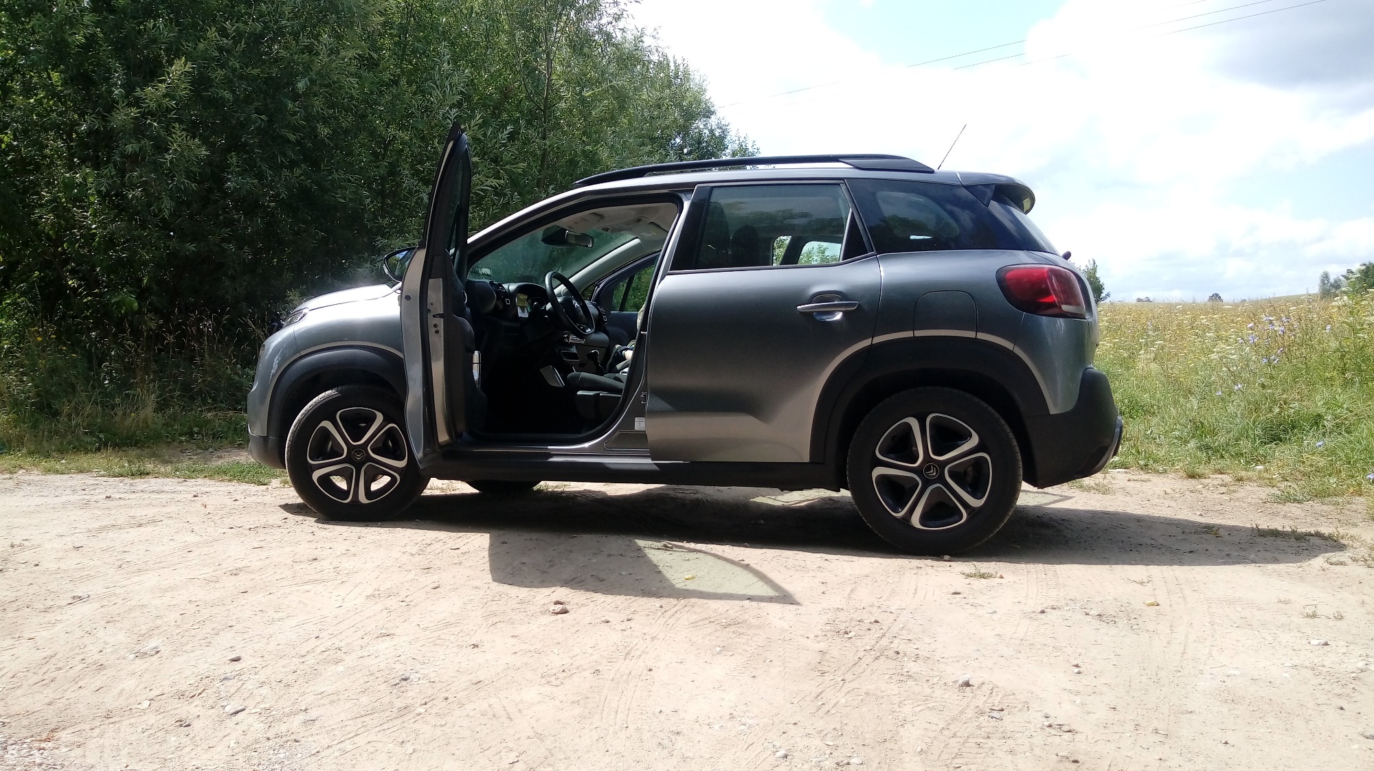 Szary samochód osobowy z otwartymi drzwiami kierowcy, stojący na polnej drodze, w tle zieleń drzew i kwitnące pole.