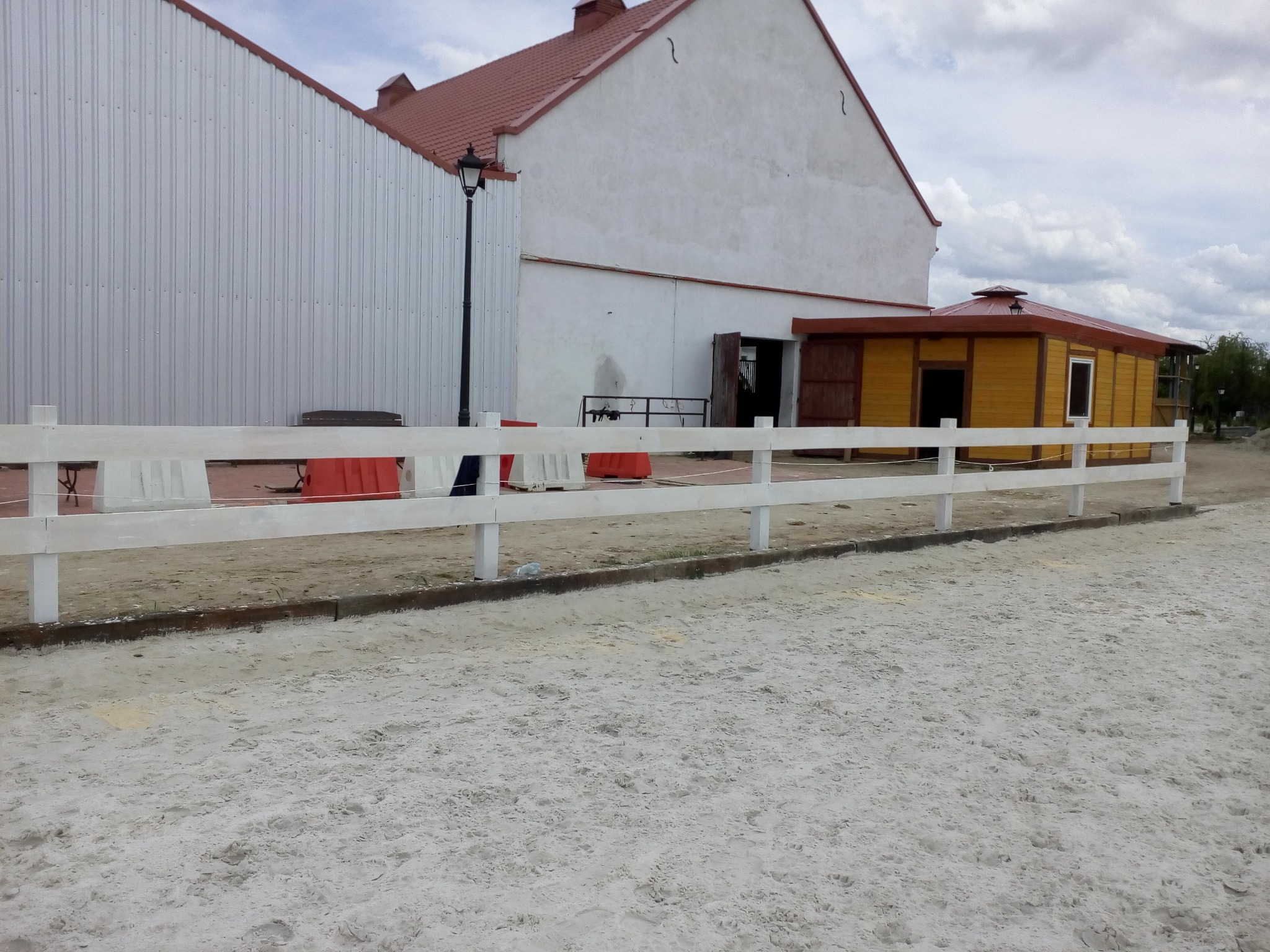 Nowoczesne, białe ogrodzenie panelowe z drewnianych desek na piaszczystym podłożu, w tle budynki gospodarcze o różnej architekturze i kolorystyce.