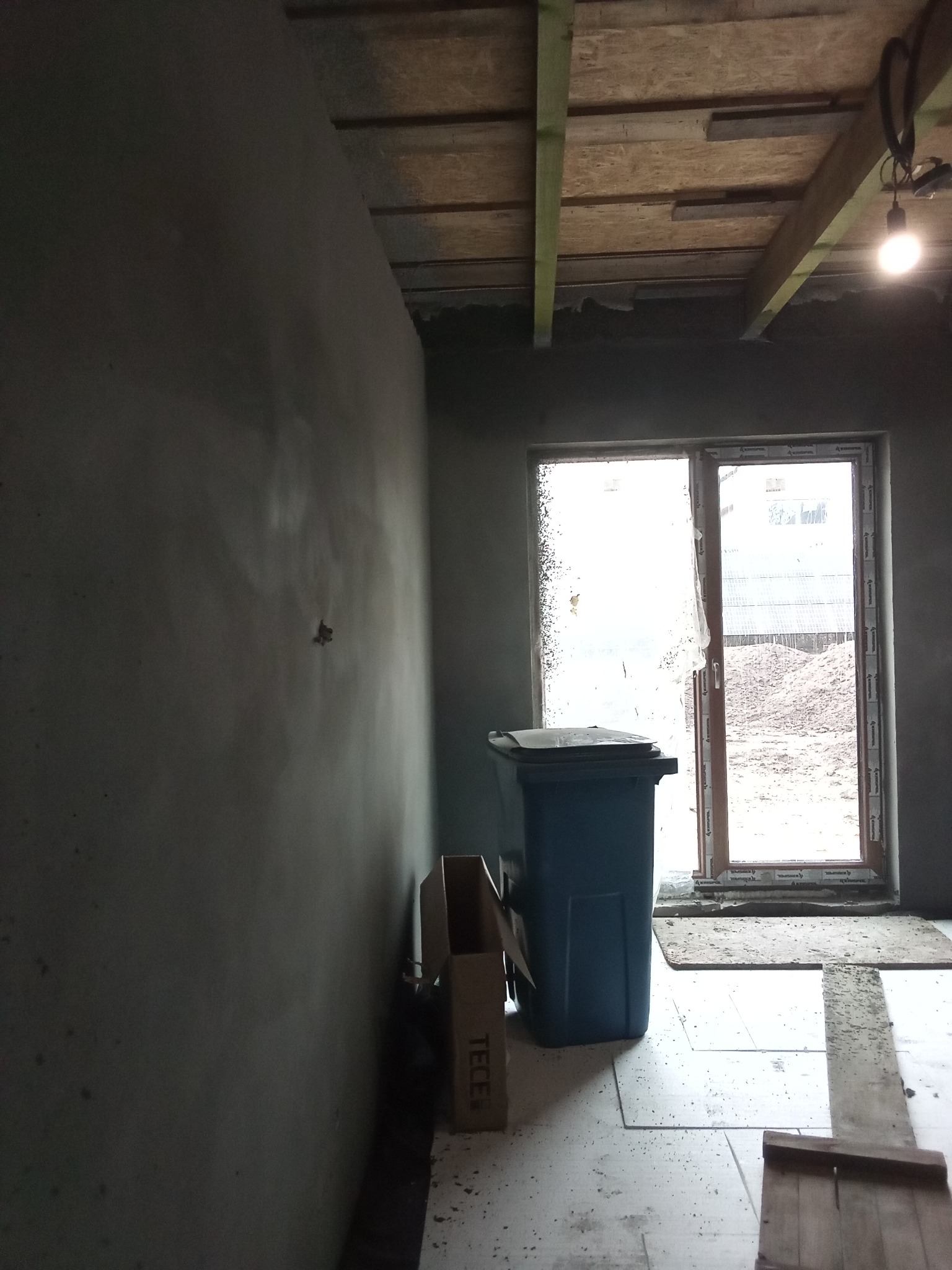 Ściana po nałożeniu tynku maszynowego, widoczny sufit z drewnianą konstrukcją, okno zabezpieczone folią, niebieski pojemnik na gruz i kartonowe opakowanie na podłodze.